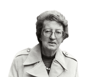 A black-and-white photograph of an elderly woman with glasses and short, wavy hair, wearing a trench coat and a pearl necklace. She stands on a city street, giving a composed expression, evoking a sense of sophistication and resilience. The urban background suggests a mid-20th-century setting.