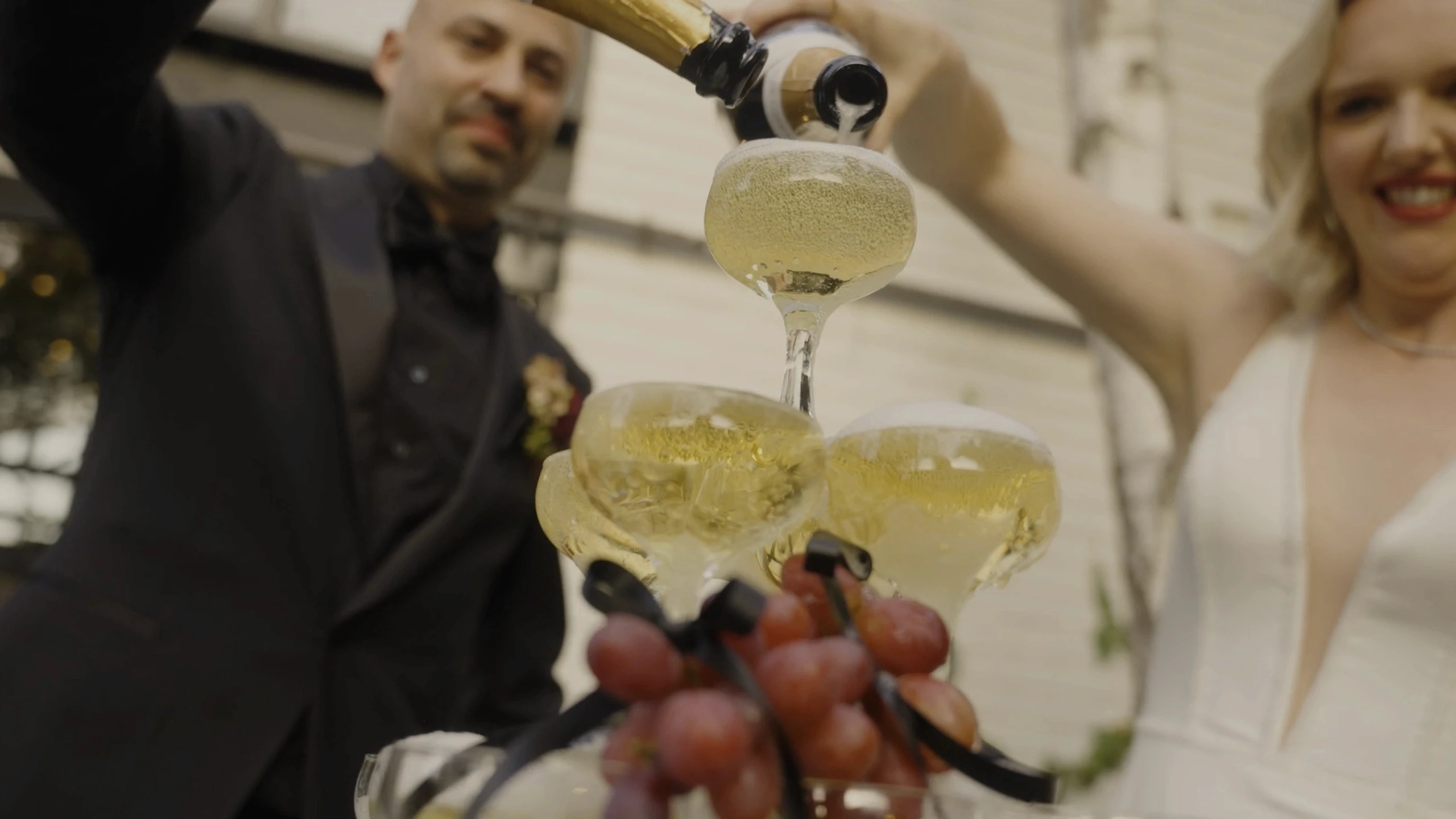 A bride and groom pour champagne into a tiered cascade of glasses, adorned with purple grapes, during a festive outdoor celebration.