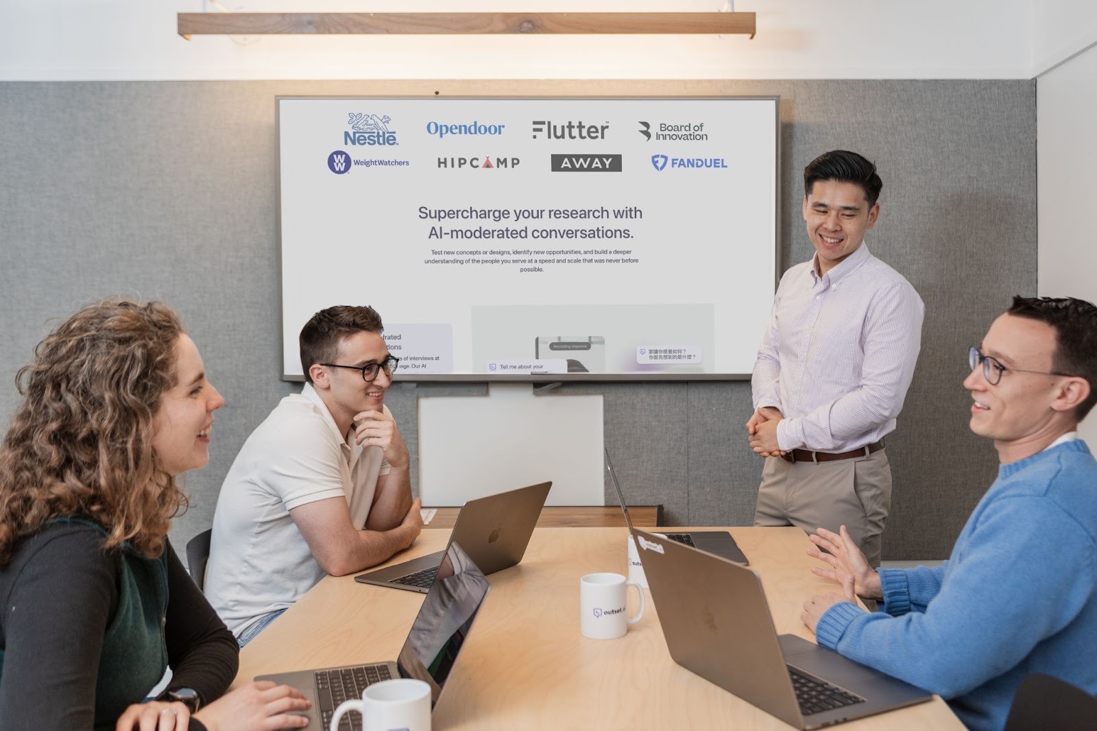 Team in a conference room discussing a presentation displayed on a large screen, with laptops and mugs on the table