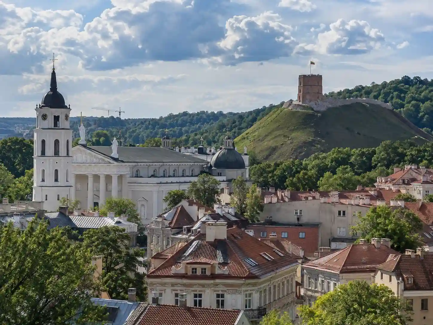 Daylight panoramic view of Vilnius Old Town in Lithuania, with Vilnius Cathedral, historic red rooftops, greenery and Gediminas Castle Tower on the hill.