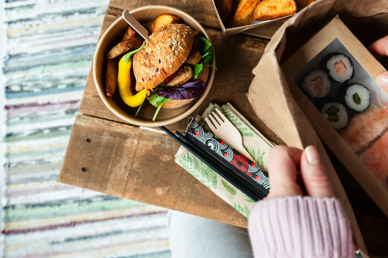 Overhead view of takeaway meal with seeded bagel sandwich, sushi box, fries, and chopsticks on wooden board.