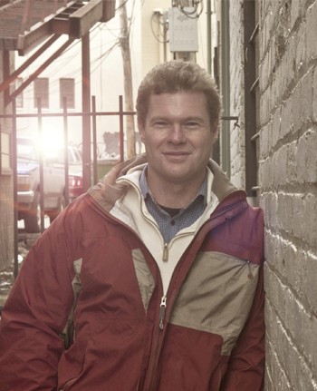 A smiling person stands against a wall in a colorful jacket, with sunlight filtering through nearby buildings.