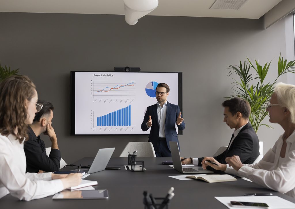 Un teamleader in piedi sta parlando ad altri dipendenti seduti ad un tavolo riunioni. Alle sue spalle un monitor su cui compare una schermata di una timesheet app