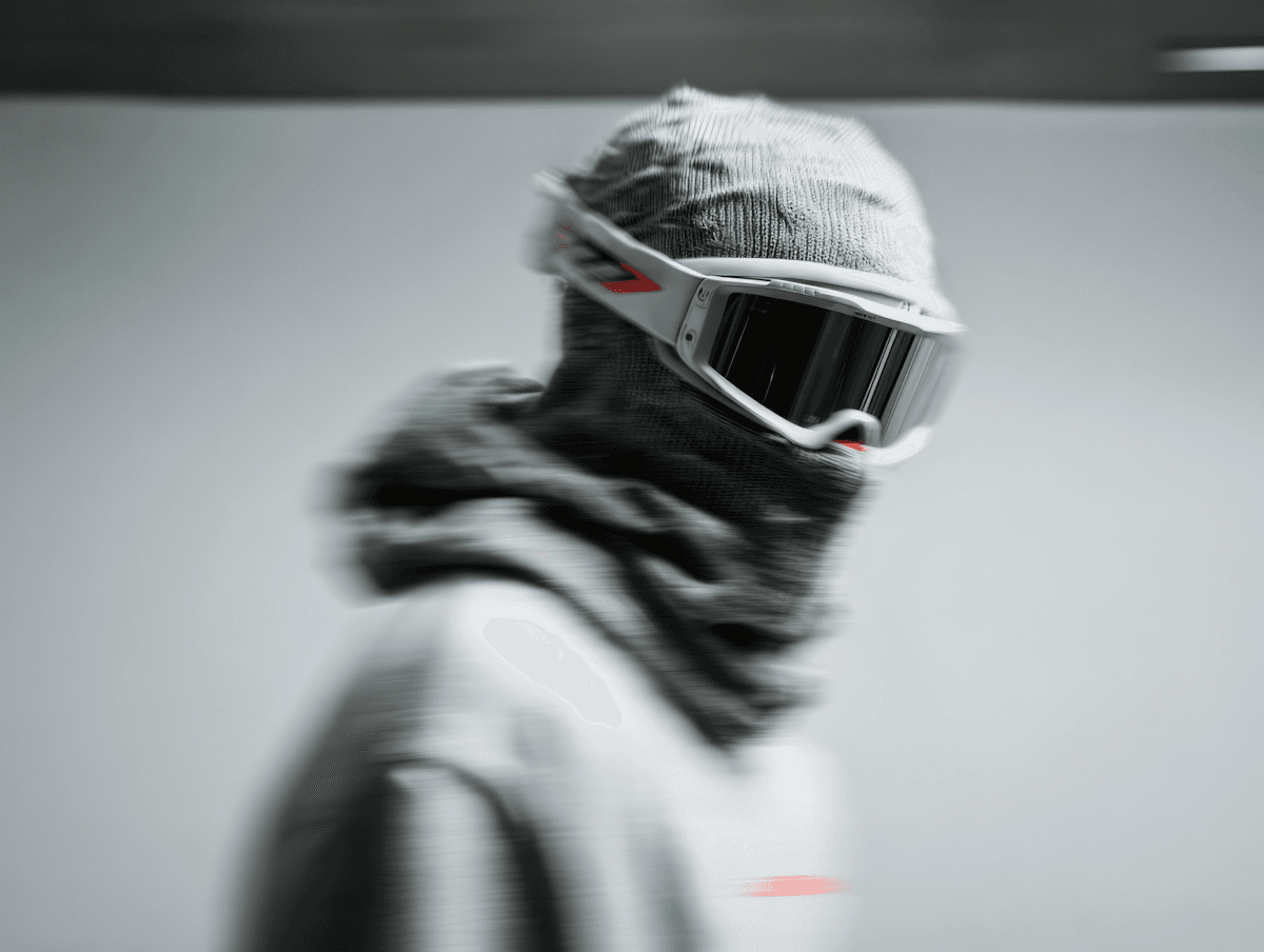 A person in a gray balaclava and white goggles with reflective lenses, set against a minimalistic background.