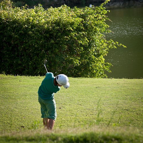 Persona con un atuendo verde jugando al golf, balanceando un palo en un campo de hierba con un lago y arbustos al fondo.