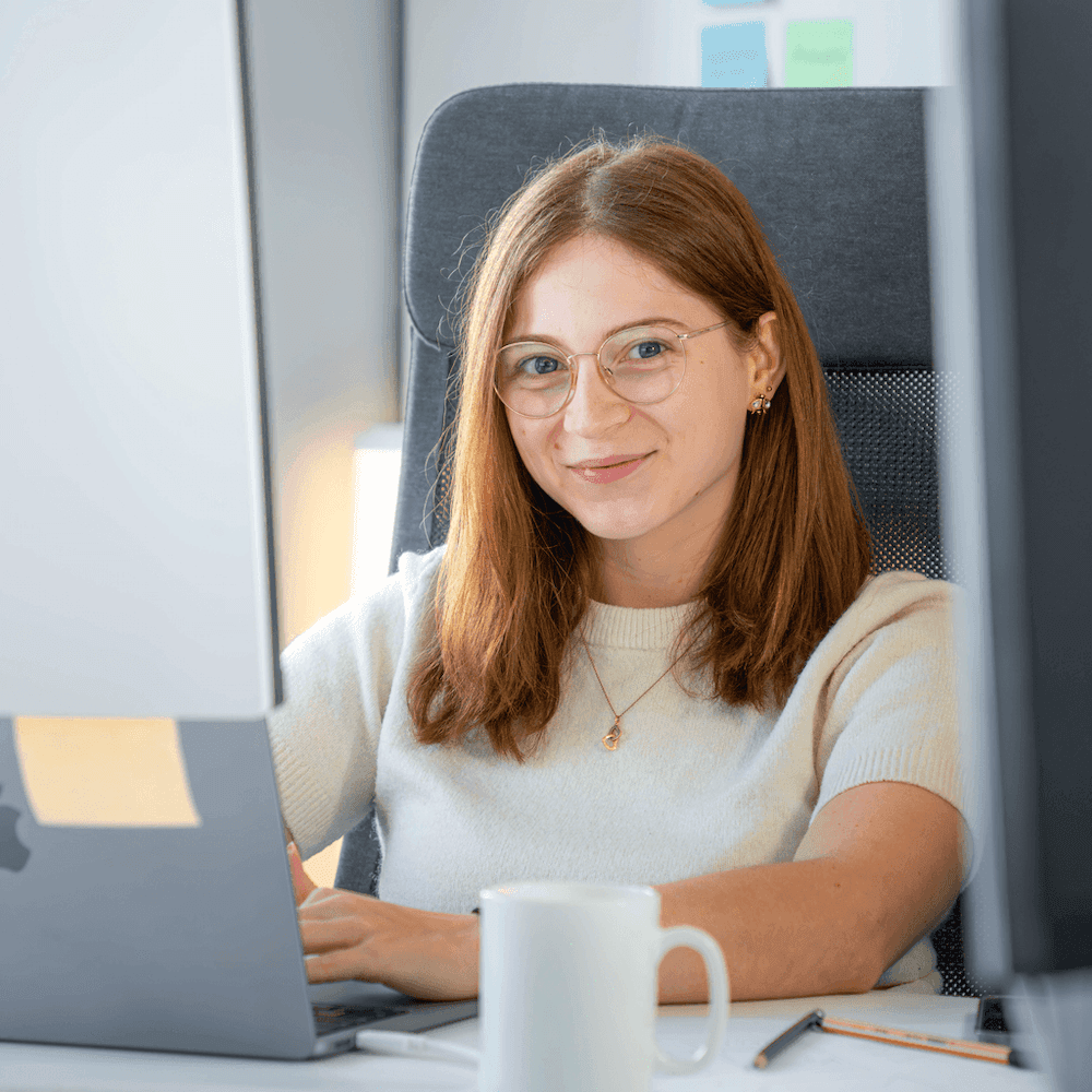 Frau mit schulterlangen braunen Haaren sitzt an einem Schreibtisch und arbeitet an einem Laptop, vor ihr steht eine weiße Tasse