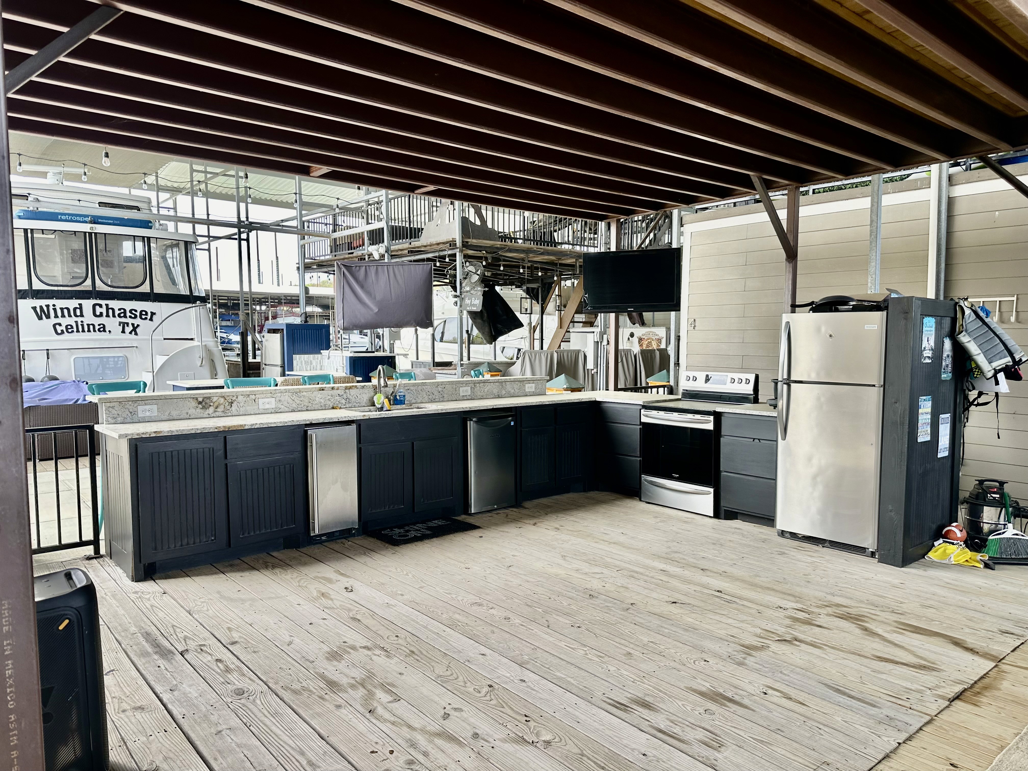 Outdoor kitchen on a spacious wooden deck featuring modern appliances, a stainless steel refrigerator, stove, and sink, with multiple TV screens and a partial view of a boat labeled "Wind Chaser" docked nearby.