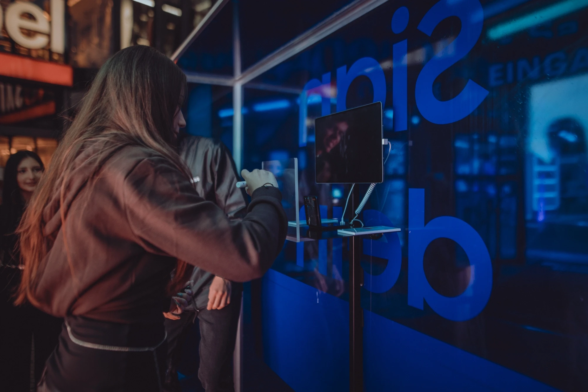 Visitor signing on a touchscreen behind the glass wall of the SAMSUNG x TIKTOK event module with blue mirrored lettering.