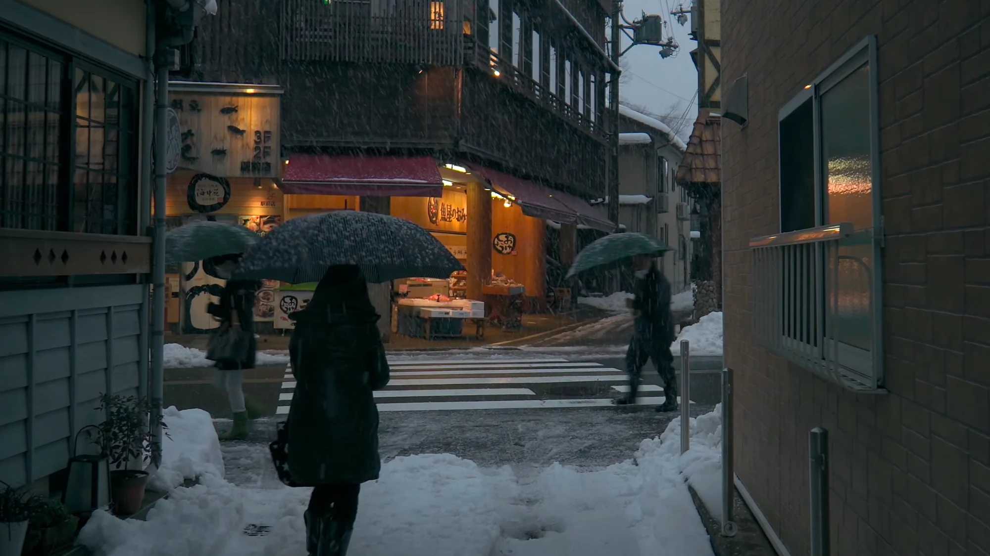 Visitors under umbrellas walking a snow-covered Kinosaki street between lit shopfronts.