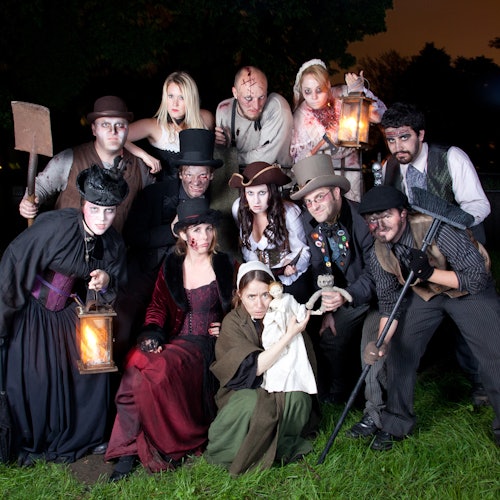 一群身穿复古服装的人手持灯笼和工具，站在草地上摆姿势，背景为黑暗的树木和橙色的天空。
