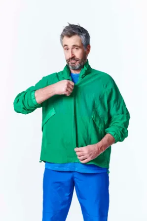 A person wearing a green jacket and blue pants adjusts their collar, with a plain light background.