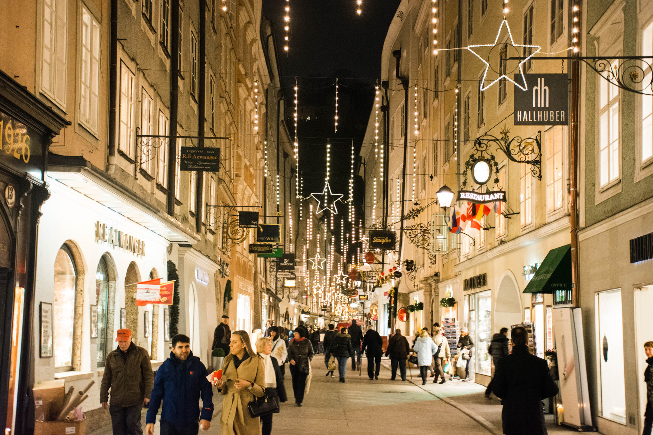 Crowds walk through a narrow European street decorated with hanging fairy lights and large illuminated stars at night.