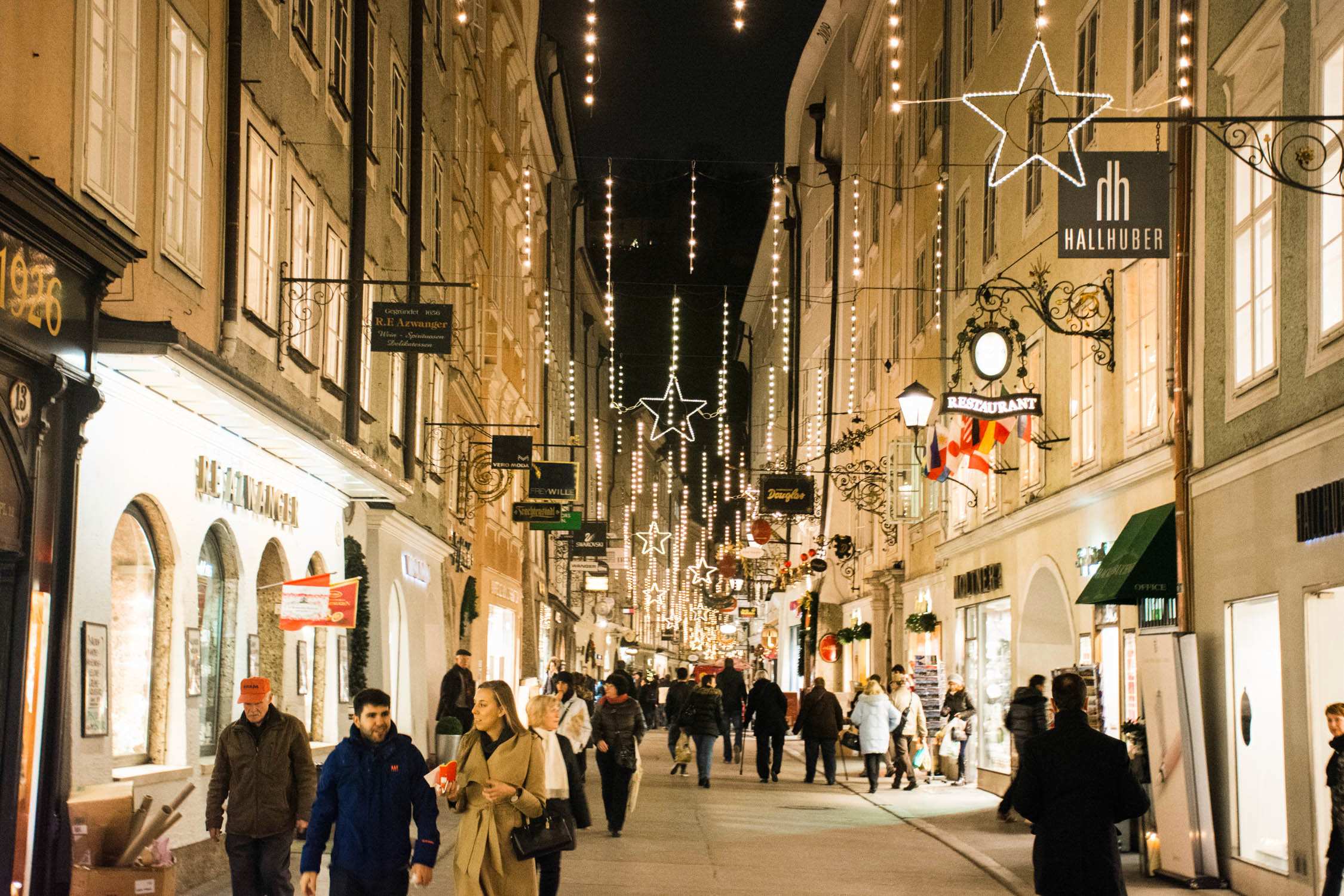 Crowds walk through a narrow European street decorated with hanging fairy lights and large illuminated stars at night.