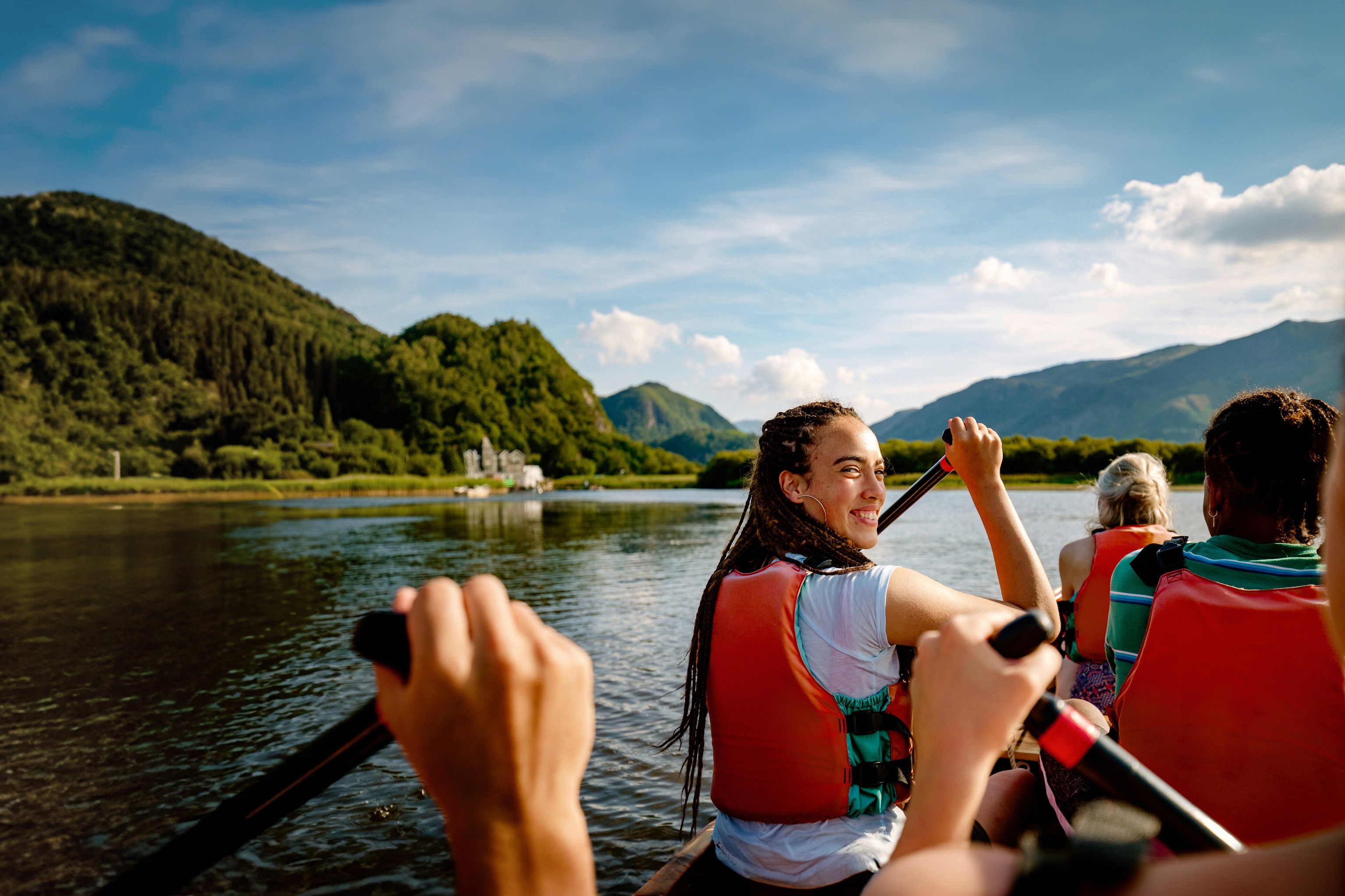Group kayaking on a calm mountain lake, paddling in life vests with green hills and distant mountains under a blue sky.