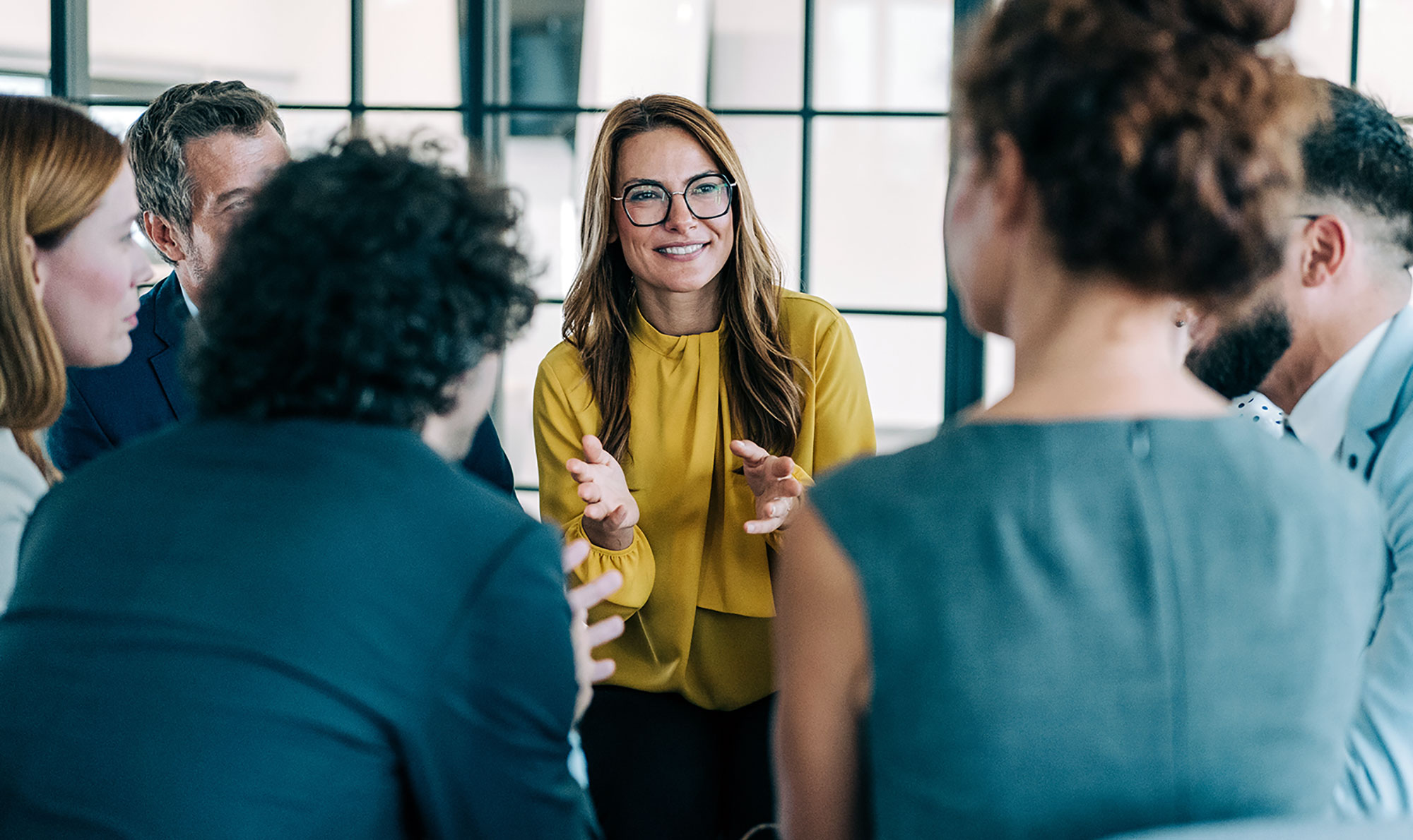 A woman wearing glasses and a mustard yellow blouse stands at the center of a circle of colleagues in a bright office with large windows. She gestures expressively while speaking to the attentive group around her, demonstrating leadership, inclusive discussion, and active workplace engagement.