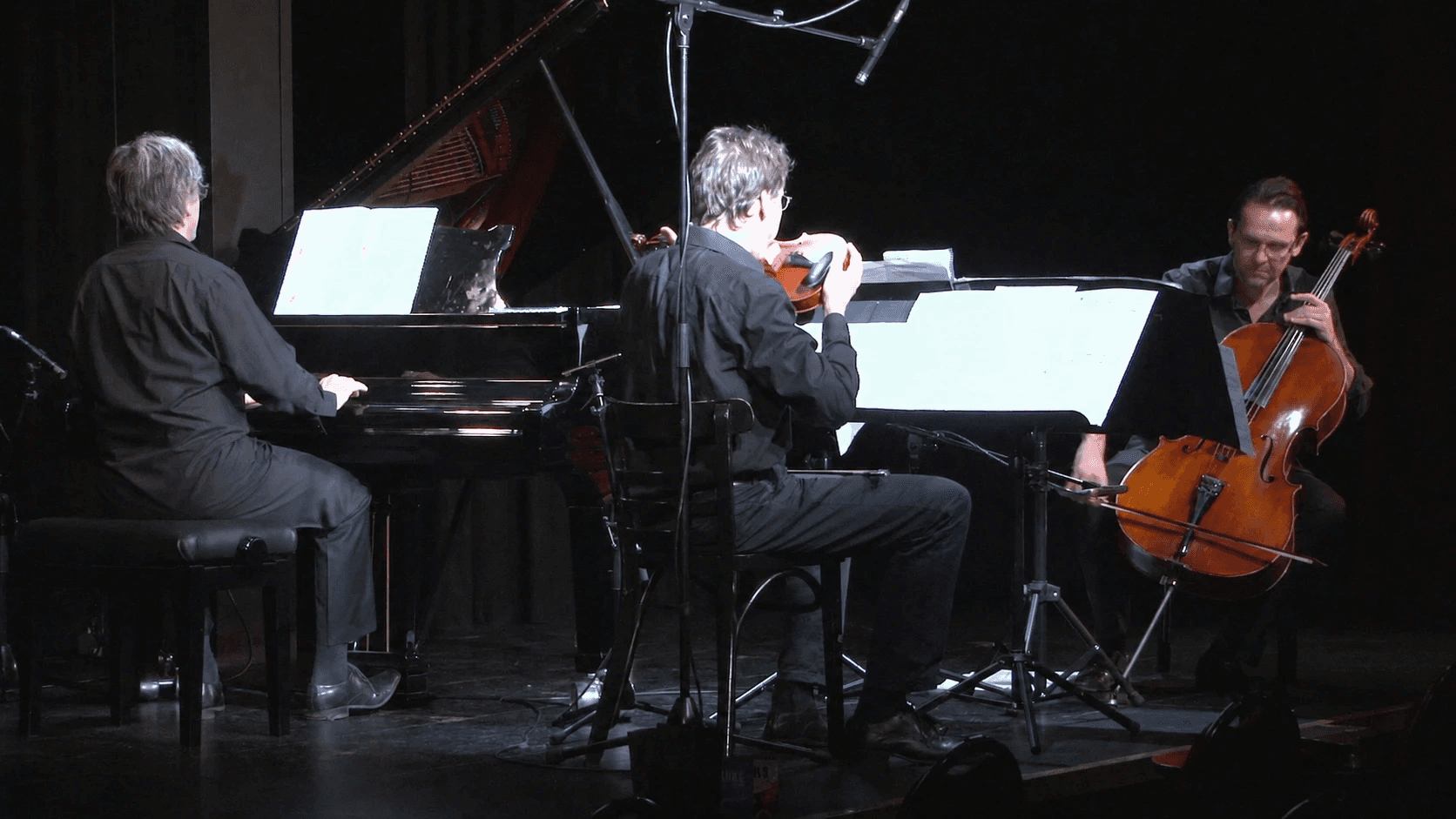 Pianist, violinist and cellist performing together on stage with music stands and amplification equipment.