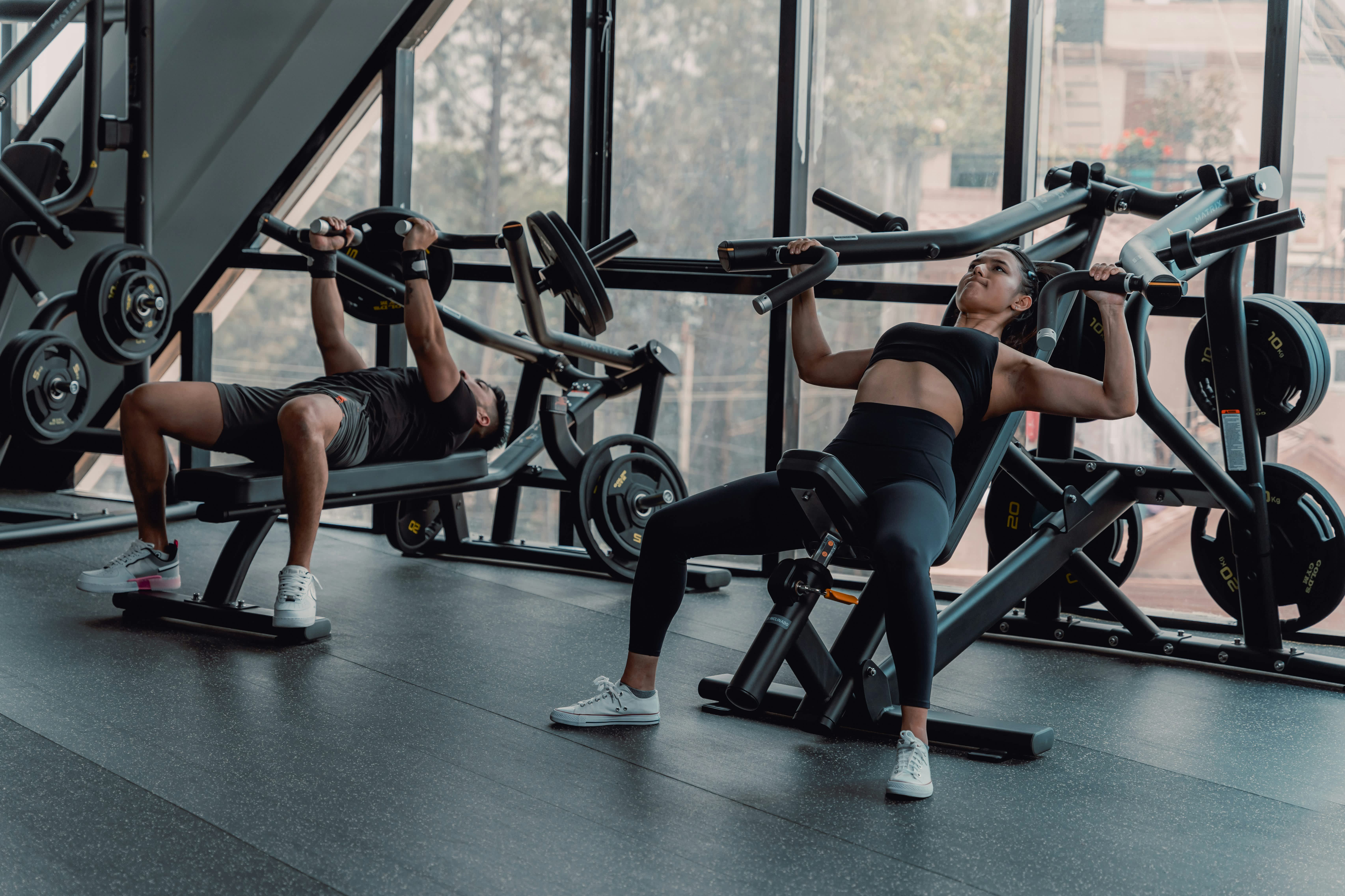 Two athletes using chest press machines in a modern gym with large windows and weight equipment.