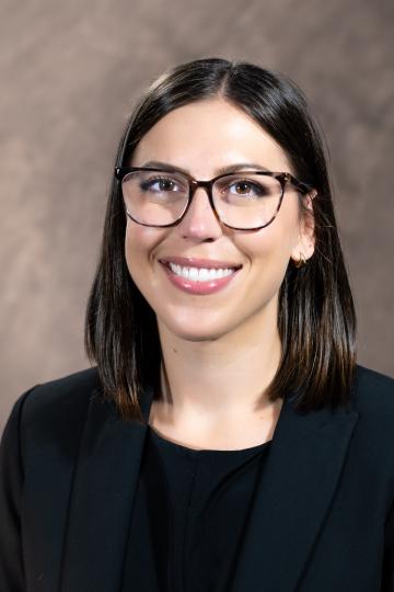 A smiling woman with glasses, dark hair, and a black blazer, looking up and to the side.