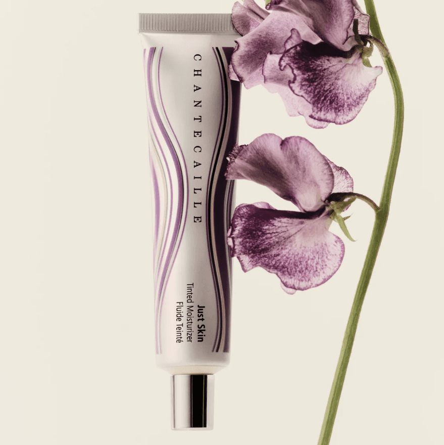 A tube of Chantecaille Just Skin Tinted Moisturizer next to a delicate purple flower on a light background.