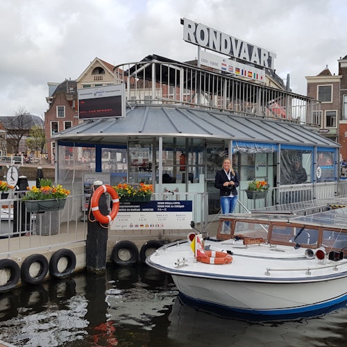 A boat docked at a quay near a building with "RONDVAART" signage. A person stands by the railing next to some flowers.