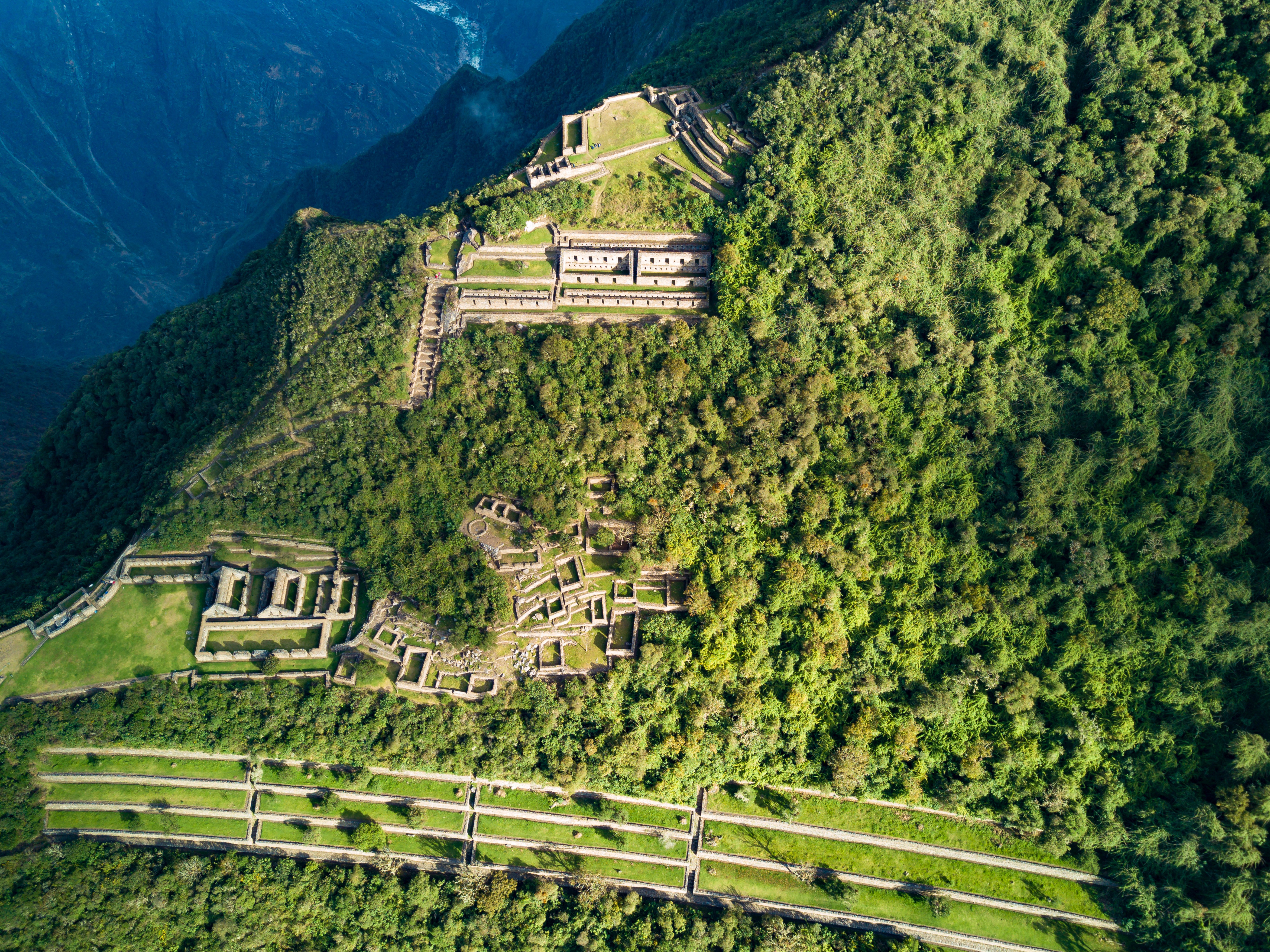 A bird's-eye view of the ancient Incan city of Choquequirao, whose ruins look like geometric shapes against the greenery that surrounds it