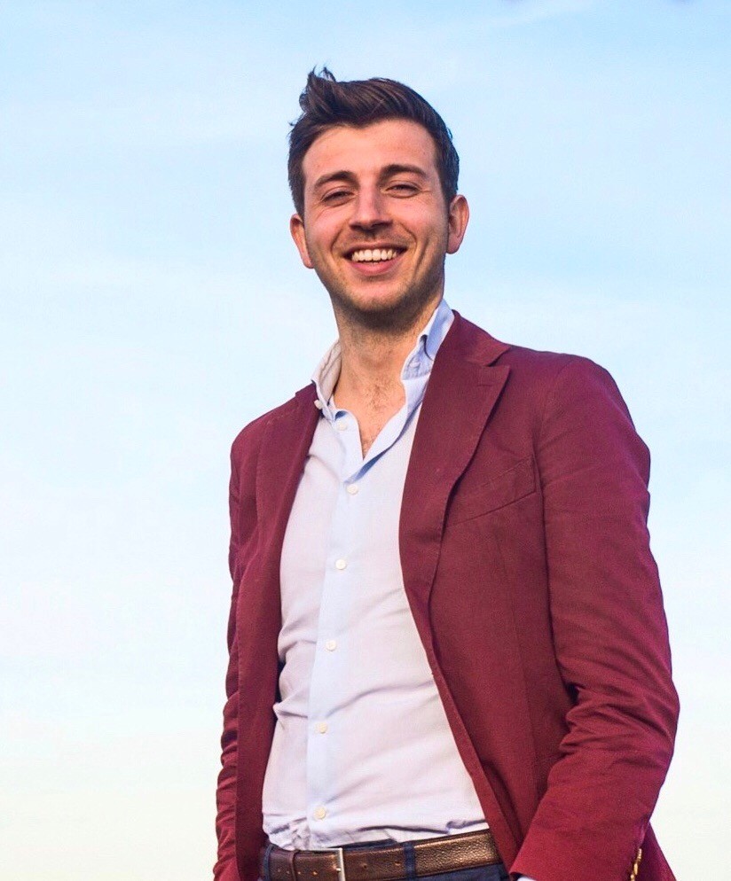 A smiling person with curly hair sits at a desk, looking at a laptop against a dark background.