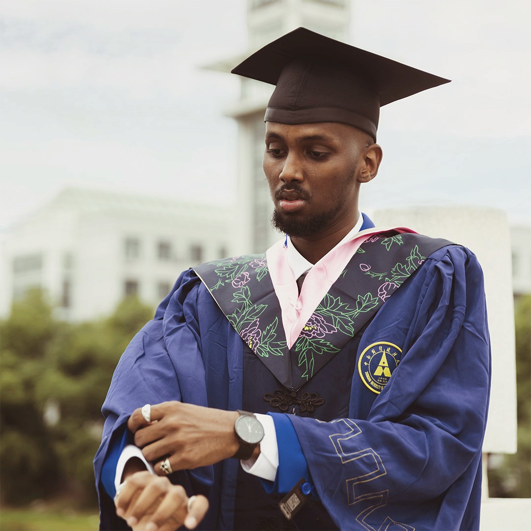 a black man wearing a purple graduation gown and a black graduation gap, he is adjusting one of his sleeves, but his expression is downcast and contemplative