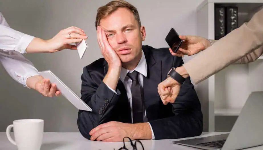 In an office setting, a weary businessman in a suit rests his face on his hand, surrounded by multiple outstretched arms holding a phone, a tablet, a document, and a watch, symbolizing overwhelming demands and stress.