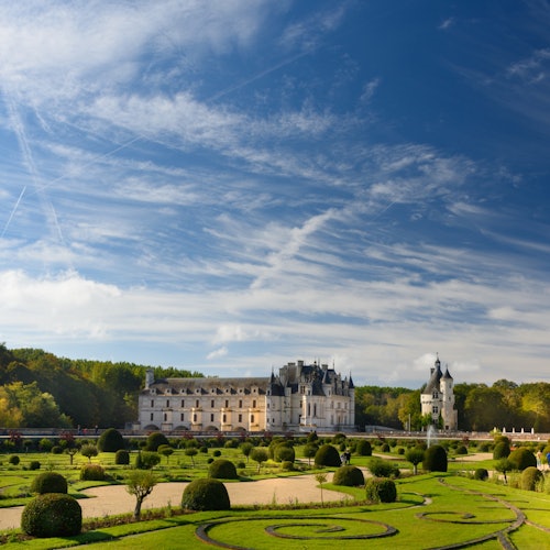 Изысканный шато с ухоженными садами и аккуратно подстриженными живыми изгородями под голубым небом с разбросанными облаками и инверсными следами.