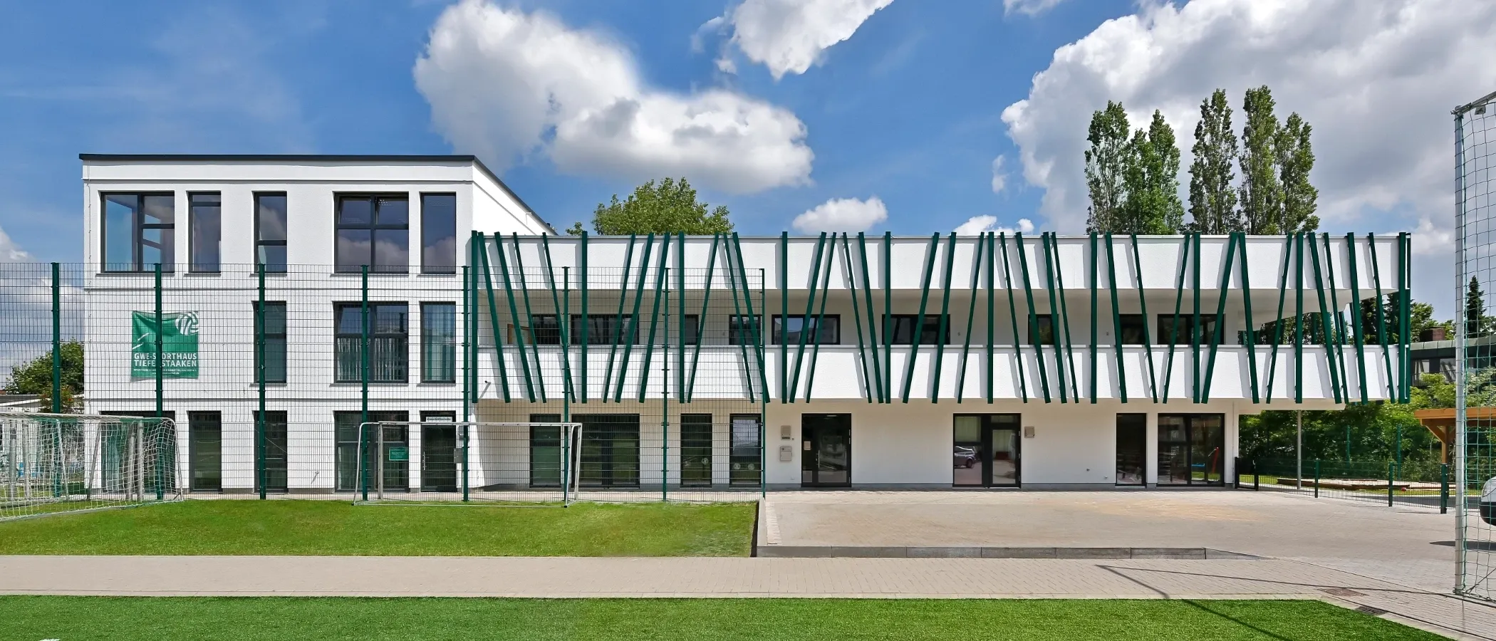Moderne Architektur eines weiß-grünen Gebäudes vor blauem Himmel mit Wolken, an ein grünes Kunstrasenfeld angrenzend.