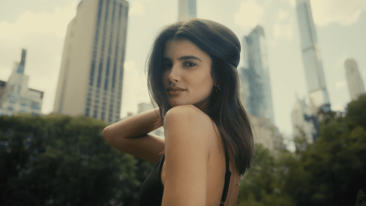A close-up of a model standing outdoors, with her hand brushing through her hair, framed by a backdrop of city buildings