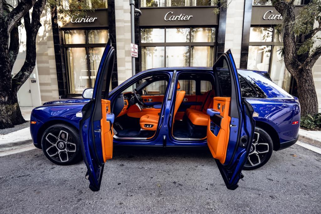 Blue Rolls-Royce Cullinan with signature coach doors open displaying orange leather interior, parked on decorative stone driveway