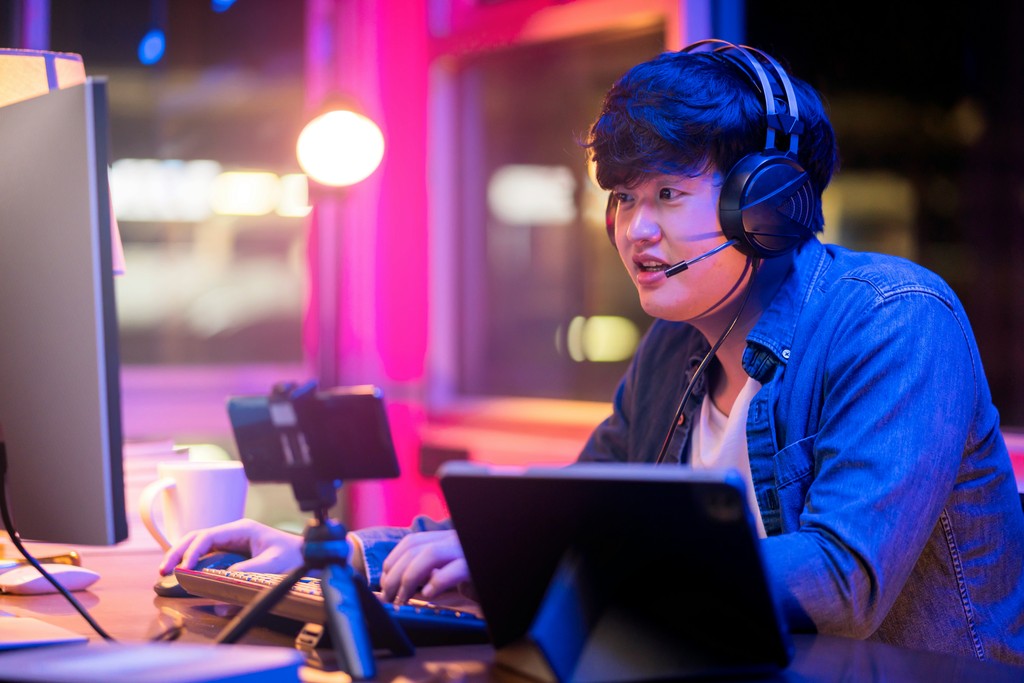 A young man with headphones and a microphone enthusiastically engages in a gaming or streaming session on his computer, surrounded by a vibrant, neon-lit environment, showcasing the immersive world of online entertainment and communication.