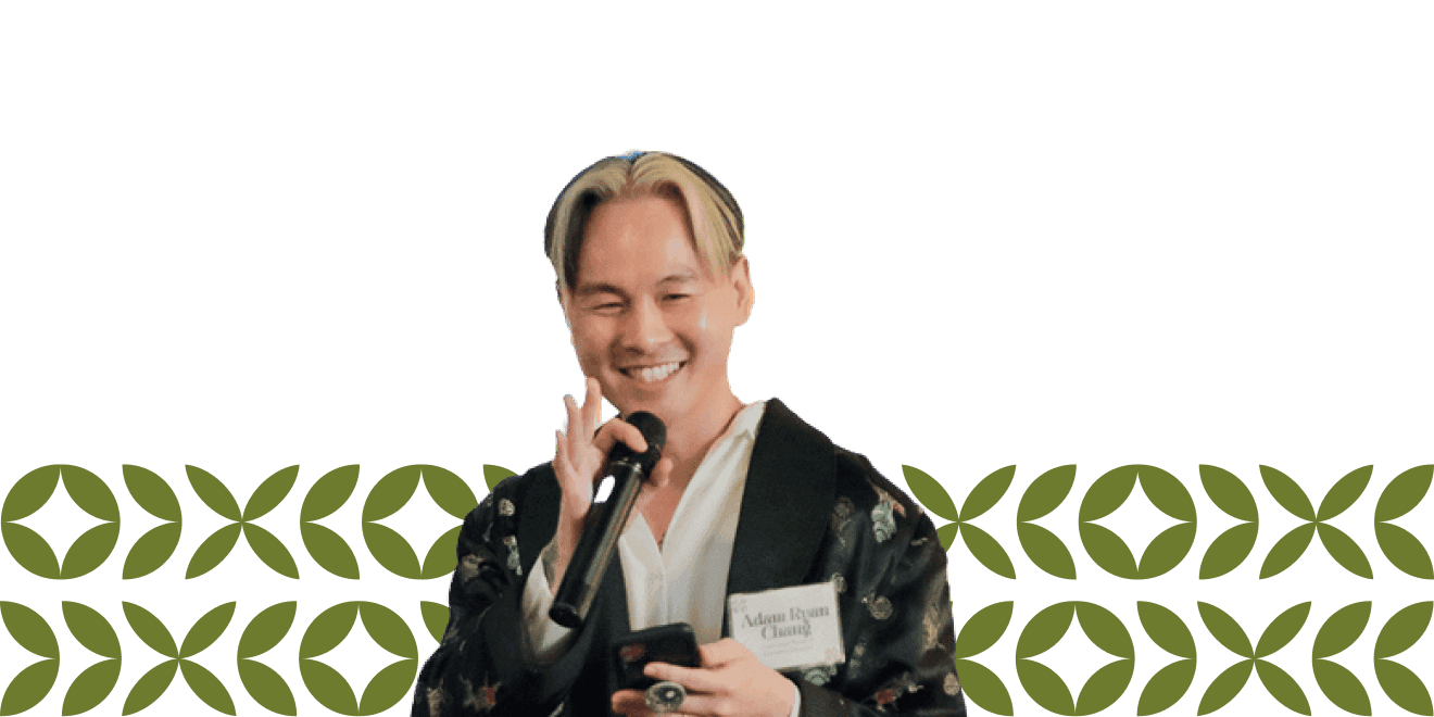 Uma pessoa segurando um microfone, sorrindo e vestindo um elegante manto preto estampado com um crachá, em pé diante de um fundo decorativo.