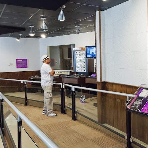 Osoba stojí v moderní výstavní místnosti a sleduje expozici elektronického vybavení, obklopenou informačními panely a obrazovkami.