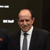 A young woman with long hair smiles while standing against a green backdrop, wearing a casual dark sweater.