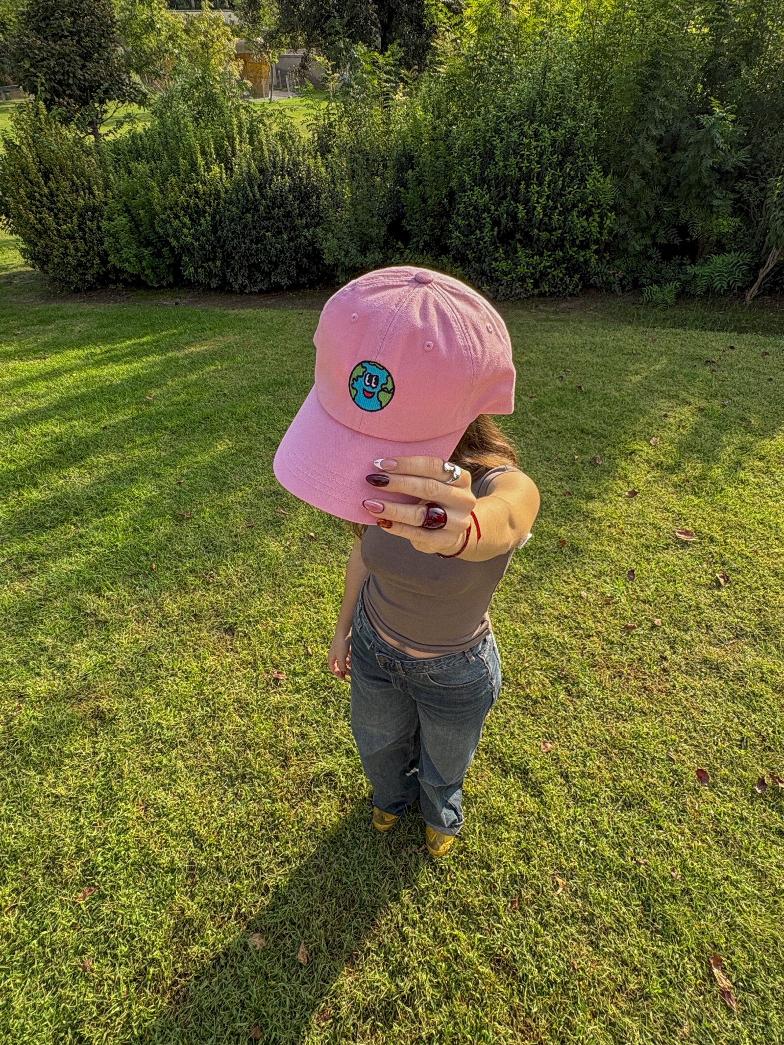 Person wearing a pink cap with a logo, standing in a grassy area with greenery.