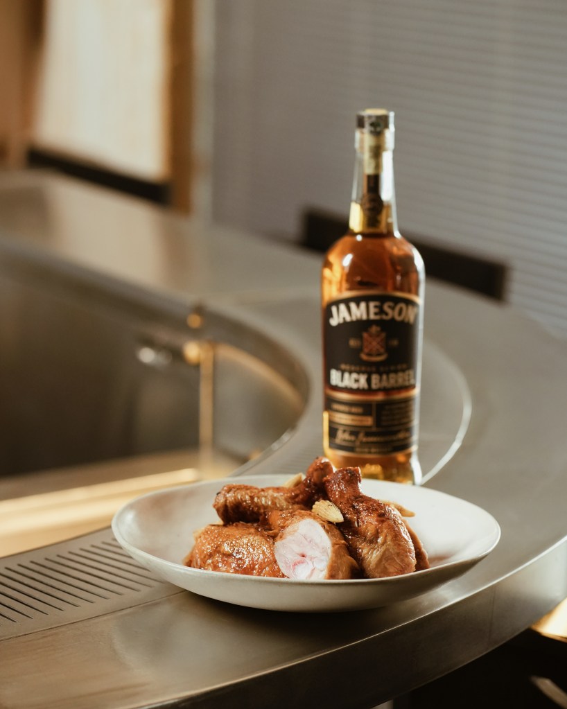 A plate of cooked chicken pieces garnished with garlic, placed next to a bottle of Jameson Black Barrel whiskey on a kitchen counter.