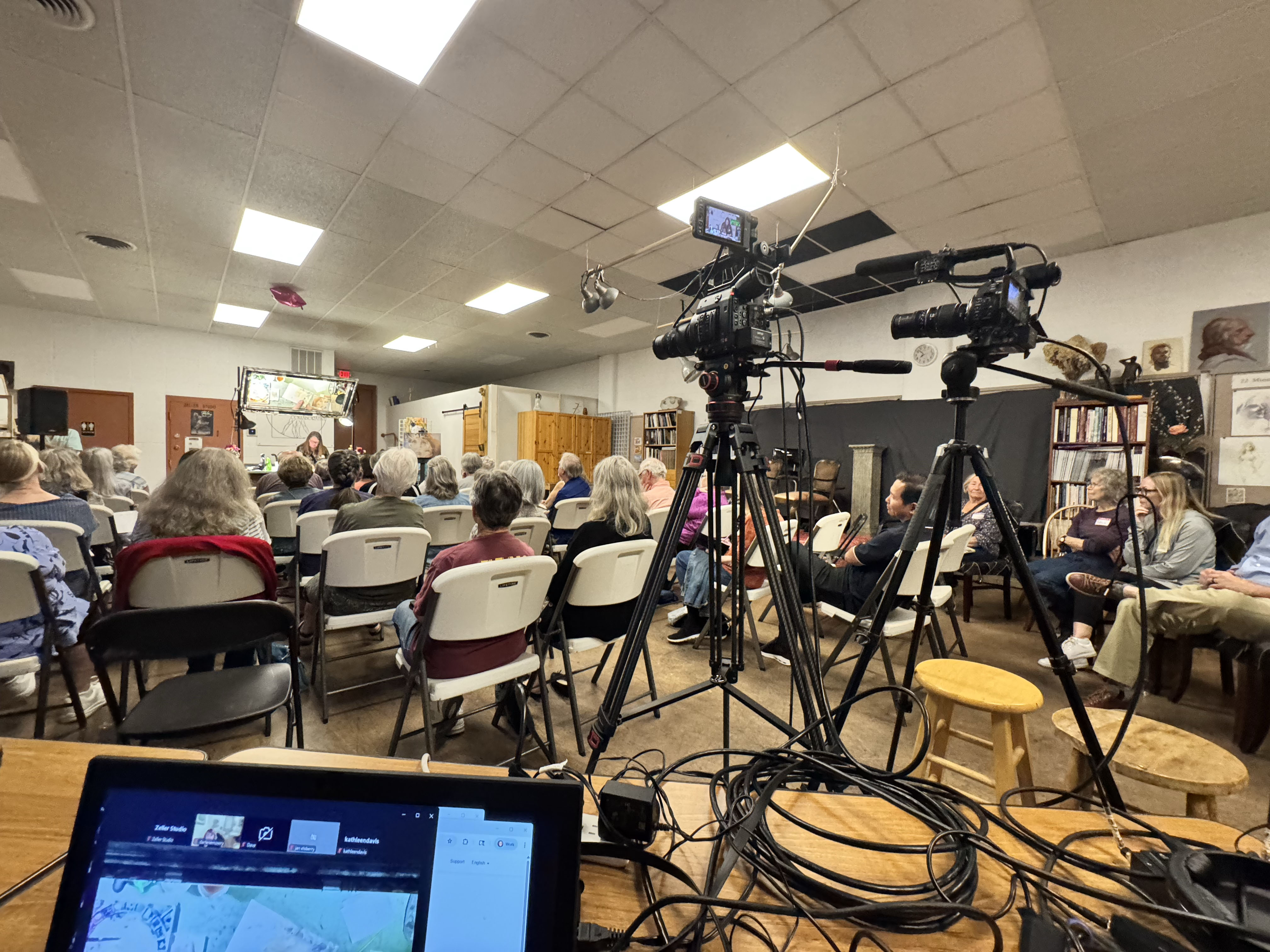 Two video cameras on tripods recording a presentation for a seated audience inside a community room.