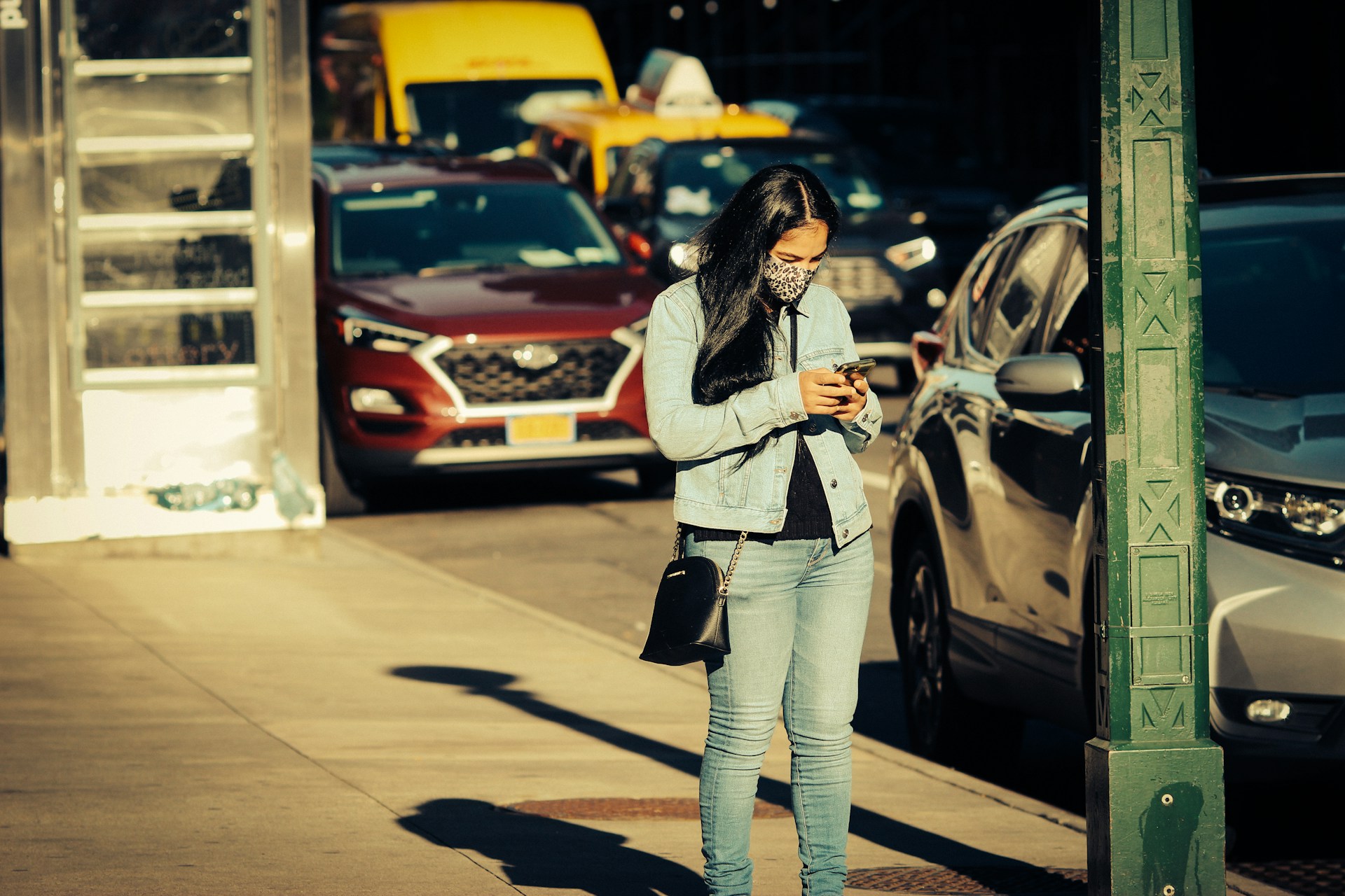 A woman standing on a city sidewalk, looking down at her phone while wearing a denim jacket and jeans, with cars and sunlight behind her.