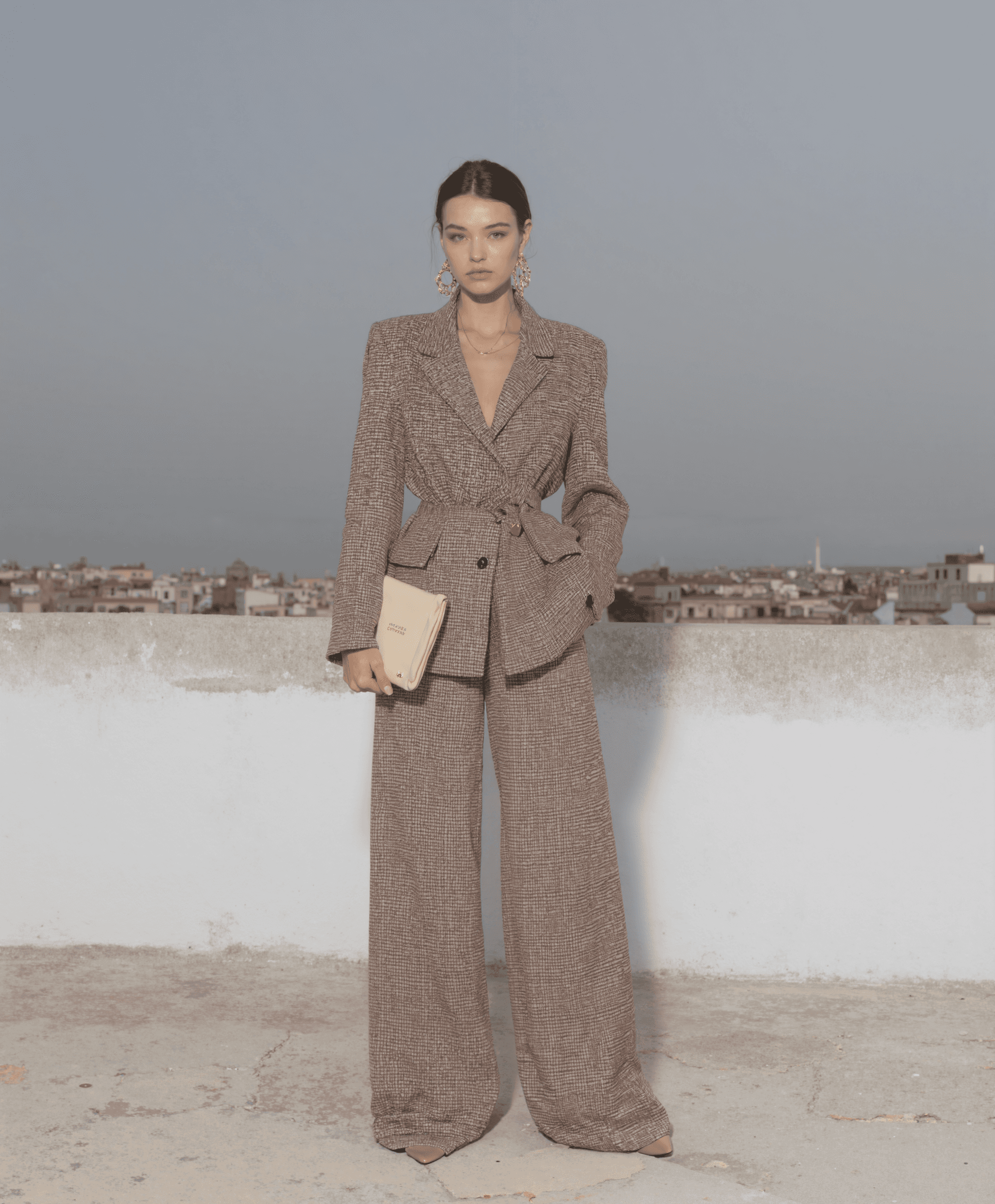 AI-generated fashion campaign visual of a woman in a belted brown suit holding a clutch on a city rooftop, produced by Myrovia Labs for La Fabrique. AI creative direction for modern womenswear brands.