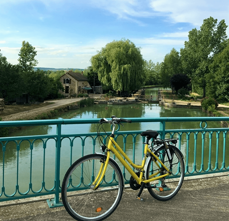 Vélo jaune appuyé contre rambarde, canal paisible et maison entourée d’arbres