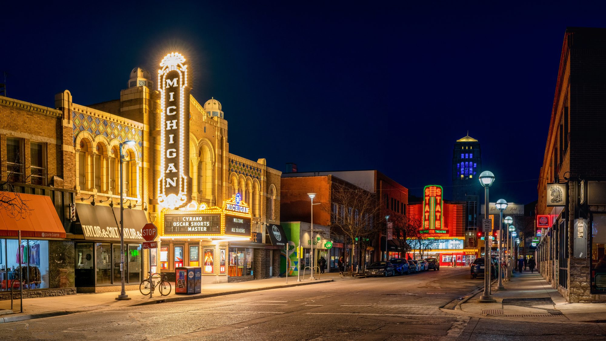 A quiet city street at night, illuminated by lights from buildings and neon signs, creating a vibrant atmosphere.