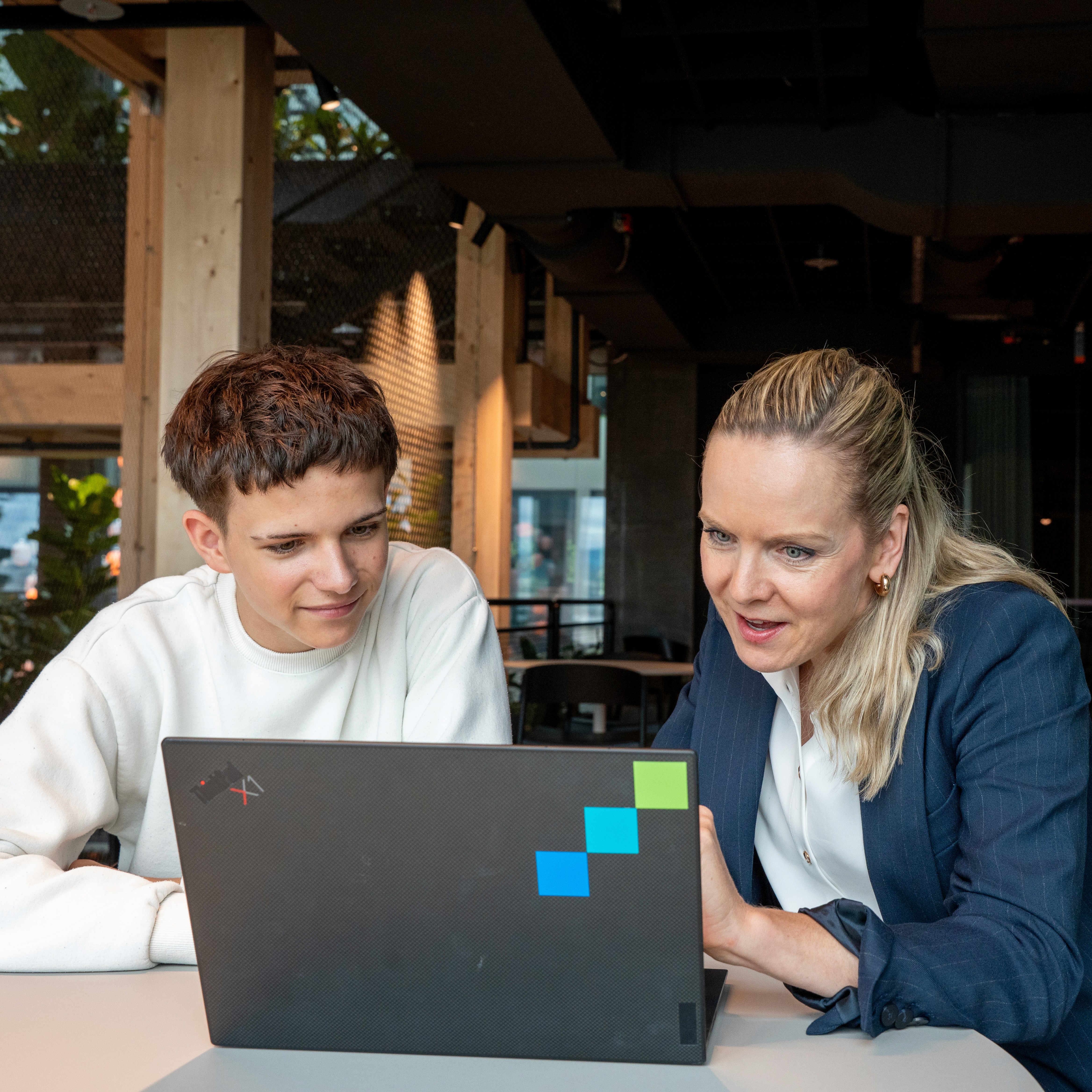 A young person with curly hair smiles at the camera while sitting in front of a laptop against a dark background.