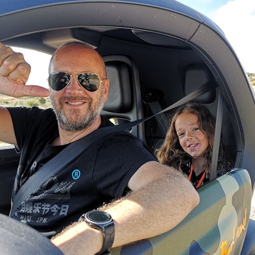 Pria tersenyum memakai kacamata hitam dan anak dengan rambut panjang di dalam mobil kecil. Pria tersebut memberikan acungan jempol; keduanya mengenakan sabuk pengaman.