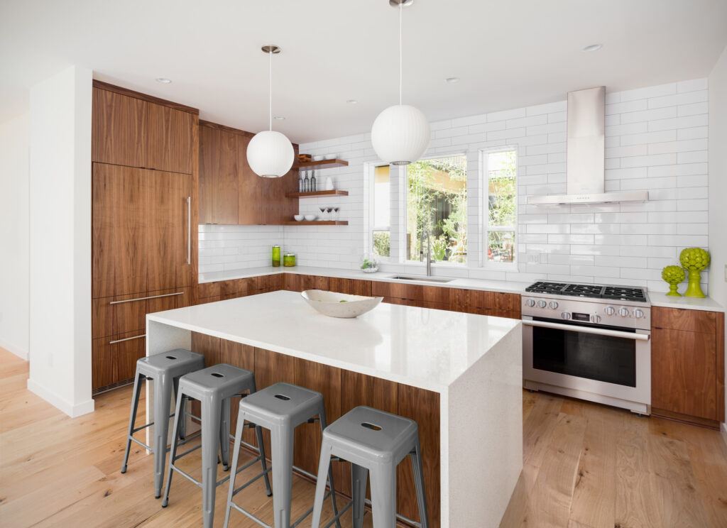 Modern kitchen with white walls and backsplash, natural wood cabinets, a white island with a waterfall countertop, and four metal barstools. Two round pendant lights hang above the island, and a stainless steel oven and hood are installed along the back wall with windows above the countertop.