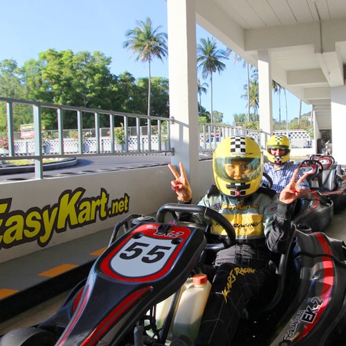 Duas pessoas em karts, com capacetes, a preparar-se para correr numa pista de karting ao ar livre, com palmeiras ao fundo.