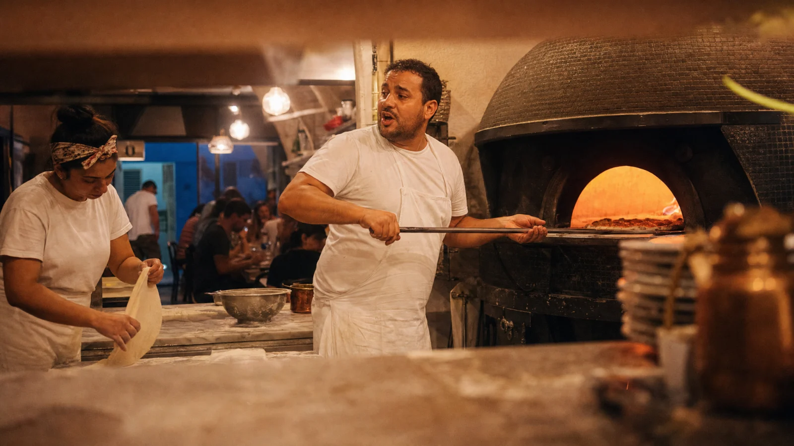 Pizzaiolo napoletano gira una pizza nel forno a legna mentre una cuoca stende l'impasto