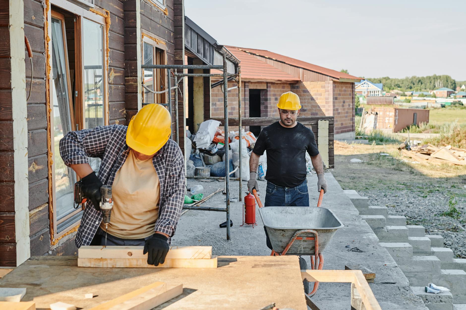 Two construction workers in yellow hard hats building a brick house with a wheelbarrow on site