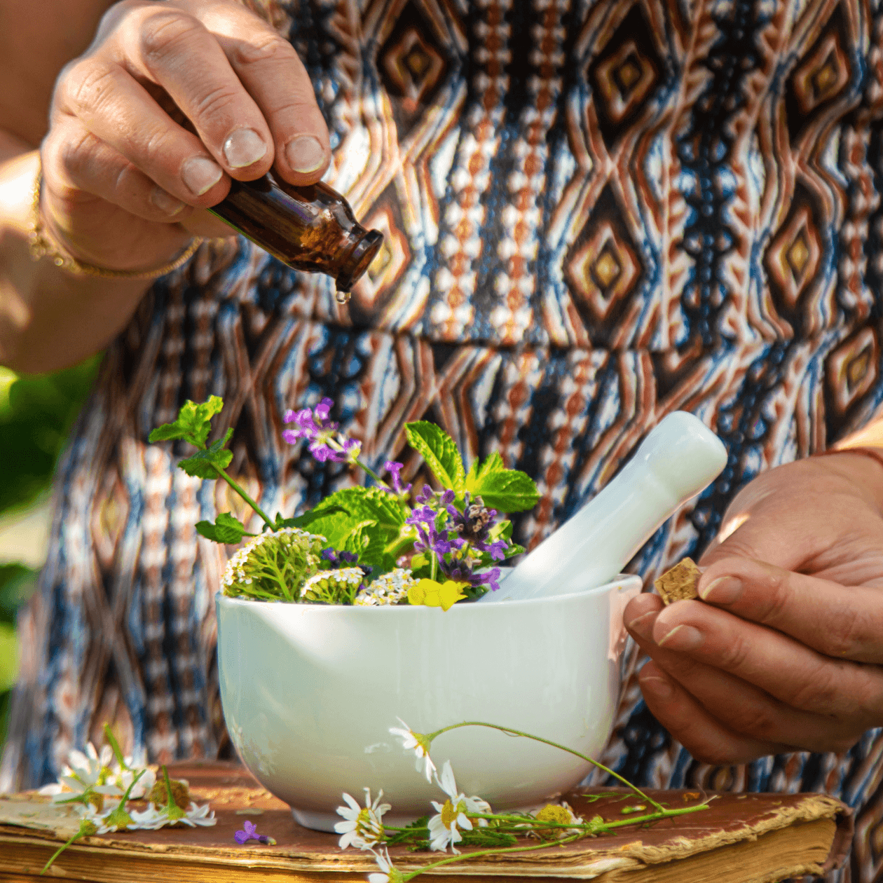 A person wearing a patterned dress prepares a traditional Ayurvedic herbal remedy, pouring liquid from a small brown bottle into a white bowl filled with fresh herbs and flowers, highlighting natural Louisville Ayurvedic services.